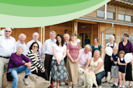 A diverse group of people, including older adults, young children, and a dog, gathered outside a modern wooden community building. Some hold balloons, while others sit on rocks or stand together, smiling. A green curved design overlays the top of the image.
